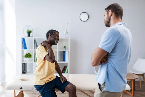 Young physiotherapist standing with crossed arms near african american patient suffering from neck pain — Stock Photo