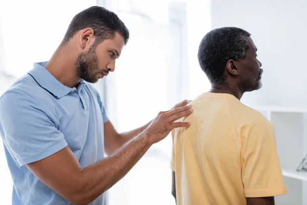 Junger Rehabilitologe untersucht Schulter und Rücken eines reifen afrikanisch-amerikanischen Mannes — Stockfoto