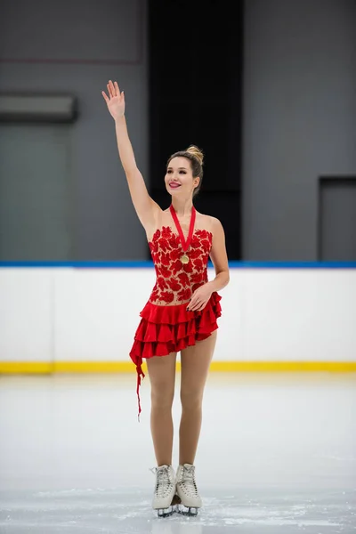 Glückliche Eiskunstläuferin in rotem Kleid und goldener Medaille winkt in Eisarena — Stockfoto