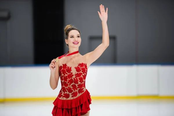Joyful figure skater in red dress holding golden medal and waving hand on ice arena — Stockfoto