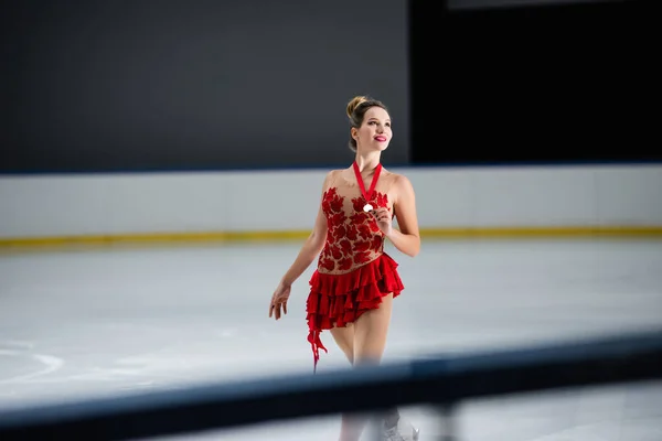 Positive figure skater in red dress holding golden medal on ice rink — Foto stock