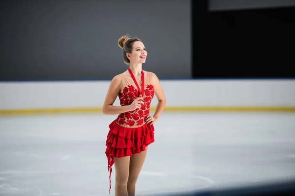 Positive figure skater in red dress holding golden medal and posing with hand on hip — Stockfoto