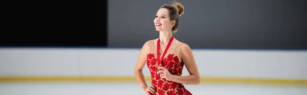 Positive figure skater in red dress holding golden medal and posing with hand on hip, banner — Stockfoto