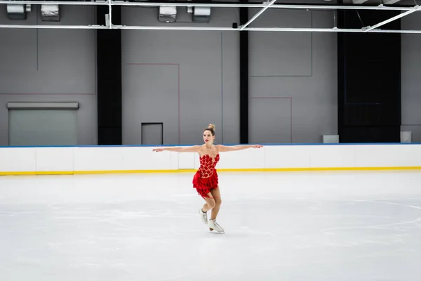 Full length of young woman in dress figure skating with outstretched hands in professional ice arena — Stockfoto