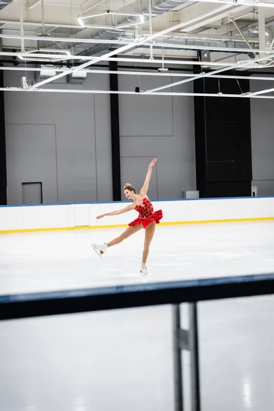 Full length of happy woman in red dress figure skating with outstretched hands in professional ice arena — Stockfoto