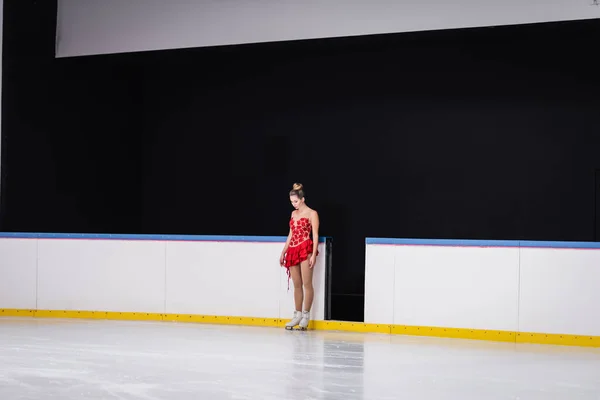 Longitud completa de joven patinador de la figura preocupado en vestido rojo de pie en pista de hielo - foto de stock