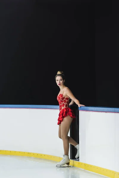 Longitud completa de joven preocupado patinador artístico en vestido rojo de pie cerca de la arena de hielo - foto de stock