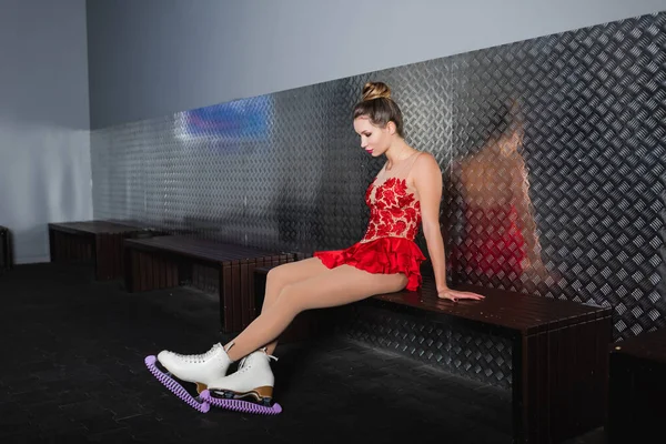 Young woman in red dress looking at figure skating shoes while sitting on bench — Stock Photo