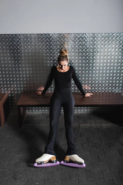 Full length of young woman in white figure skates stretching near bench — Stock Photo