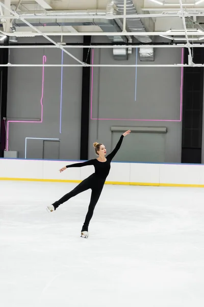 Full length of young woman in black bodysuit gesturing while skating in professional ice arena — Stock Photo