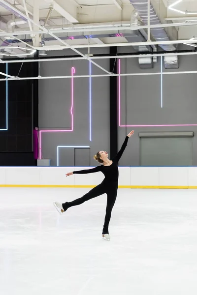 Full length of woman in black bodysuit with outstretched hands figure skating in professional ice arena — Stock Photo