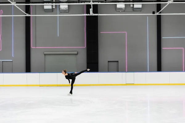 Vista lateral da jovem mulher em macacão preto patinação na arena de gelo profissional — Fotografia de Stock