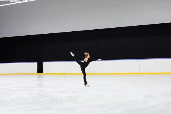 Side view of figure skater in black bodysuit skating with outstretched hand in professional ice arena — Stock Photo