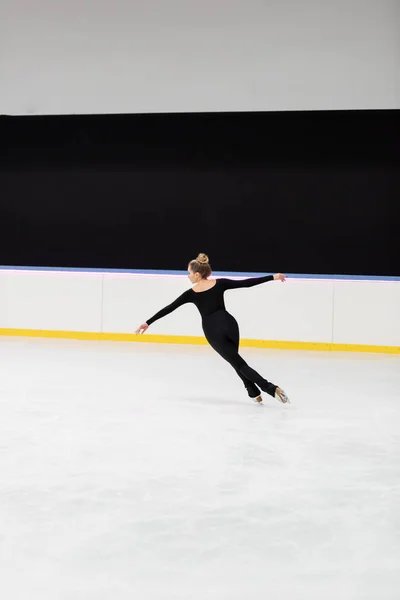 Full length of professional figure skater in black bodysuit skating with outstretched hands in ice arena — Stock Photo