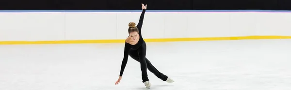 Full length of professional figure skater in black bodysuit skating with outstretched hands in ice arena, banner — Stock Photo