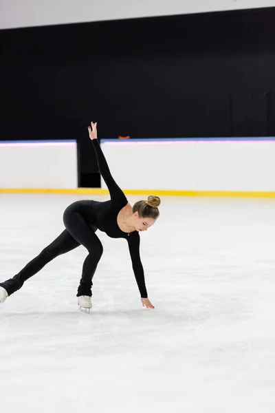 Full length of professional figure skater in black bodysuit skating with outstretched hand and touching frozen ice in arena — Stock Photo