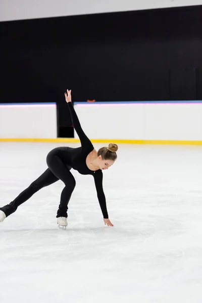 Longitud completa de patinador artístico profesional en negro bodysuit patinaje con la mano extendida y tocar hielo congelado en la arena - foto de stock