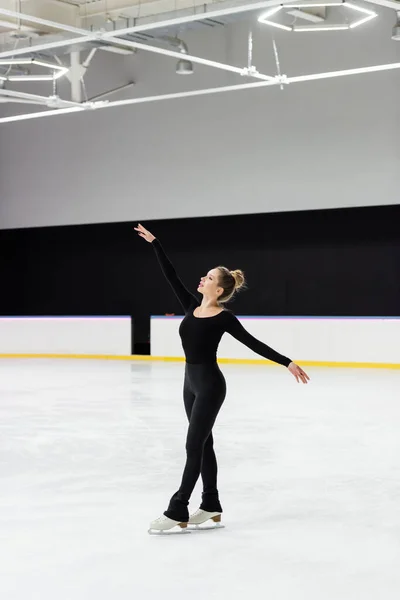 Full length of sensual figure skater in black bodysuit skating with outstretched hands in ice arena — Stockfoto