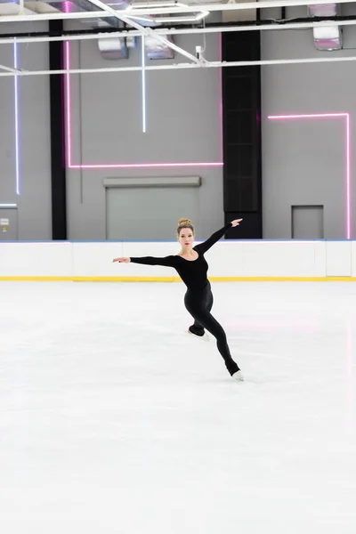 Full length of professional figure skater in black bodysuit skating with outstretched hands in ice arena with neon lights — Stockfoto
