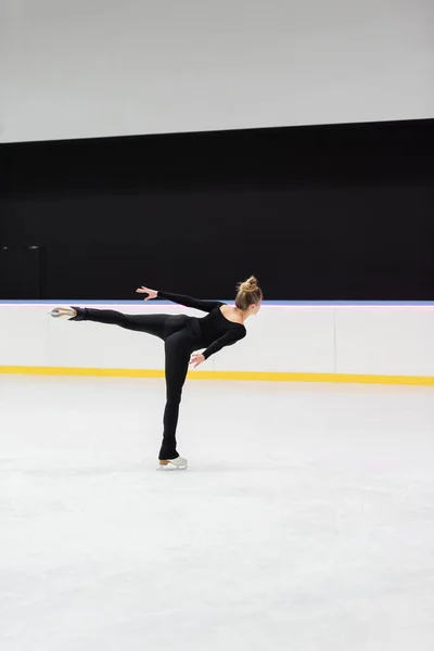 Back view of professional figure skater in black bodysuit skating with outstretched hand in ice arena — Stockfoto