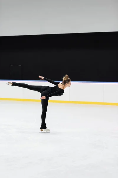 Side view of professional figure skater in black bodysuit skating with outstretched hand in ice rink — Stock Photo