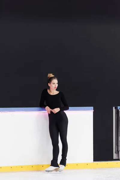 Longitud completa del joven patinador artístico en traje negro de pie sobre patines de hielo cerca de la arena de hielo congelado - foto de stock