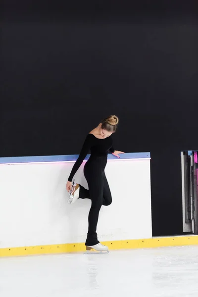 Full length of young figure skater in black bodysuit checking blade on ice skates near frozen ice arena — Stock Photo