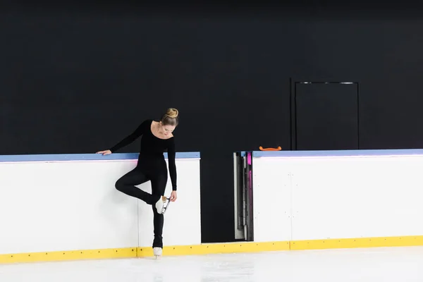 Full length of figure skater in black bodysuit checking blade on ice skates near frozen ice arena — Stock Photo