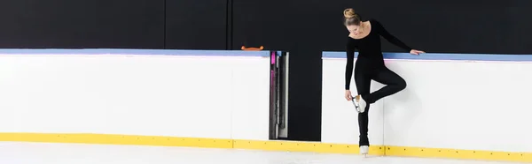 Full length of figure skater in black bodysuit checking blade on ice skates near frozen ice arena, banner — Stock Photo