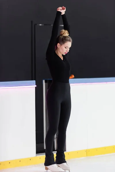 Longitud completa del joven patinador artístico en traje negro y patines de hielo que se extienden cerca de la arena de hielo congelado - foto de stock