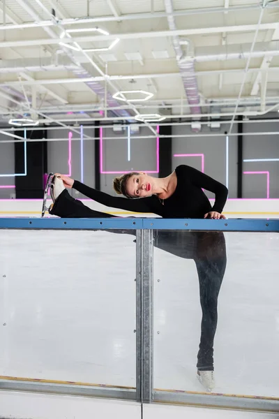 Full length of professional figure skater in black bodysuit and ice skates stretching near frozen ice arena — Stock Photo