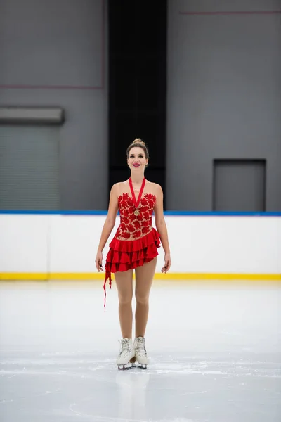 Full length of cheerful figure skater in red dress with golden medal skating on ice rink — Stock Photo