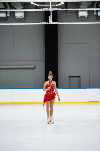 Toute la longueur du jeune patineur artistique en robe rouge regardant la médaille d'or sur la patinoire — Photo de stock