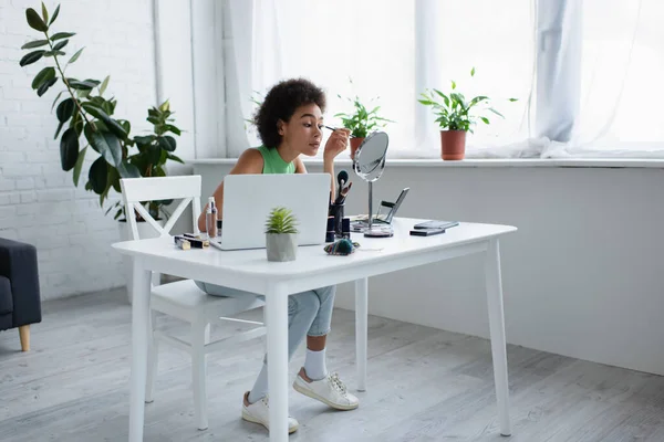 Mujer afroamericana aplicando rímel cerca de la computadora portátil en casa - foto de stock