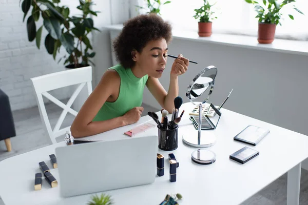 Jovem afro-americana aplicando sombra nos olhos perto de cosméticos e laptop em casa — Fotografia de Stock