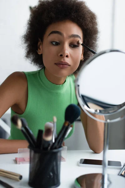 Afroamerikanerin trägt Lidschatten in der Nähe von Spiegel und Smartphone auf — Stockfoto