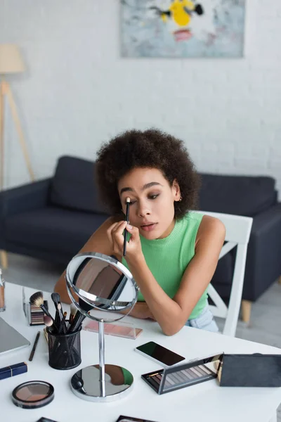 Afroamerikanerin legt Lidschatten in der Nähe von Geräten und Kosmetika auf den Tisch — Stockfoto