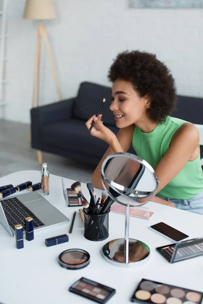Lächelnde Afroamerikanerin hält Kosmetikpinsel in der Nähe von Gadgets und dekorativer Kosmetik zu Hause — Stockfoto