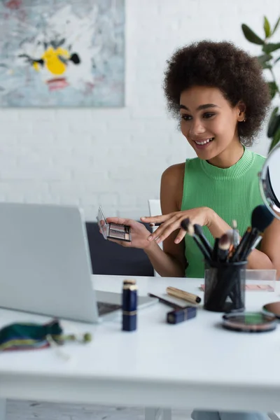 Sonriente mujer afroamericana sosteniendo cosméticos cerca de la computadora portátil borrosa en casa - foto de stock