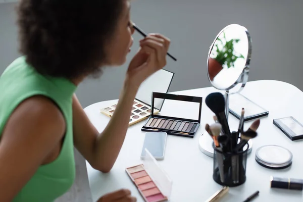 Vista lateral de mujer afroamericana borrosa aplicando cosméticos cerca del teléfono celular con pantalla en blanco y espejo - foto de stock