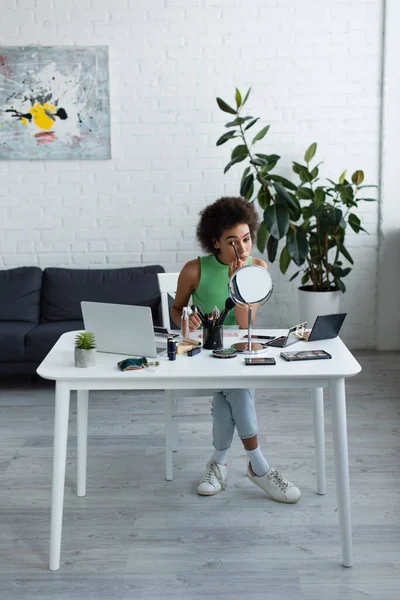 Donna afroamericana che applica cosmetici decorativi vicino a specchio e laptop a casa — Foto stock