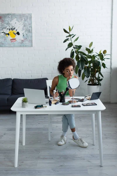 Africano americano mulher aplicando cosméticos decorativos perto de espelho e laptop em casa — Fotografia de Stock