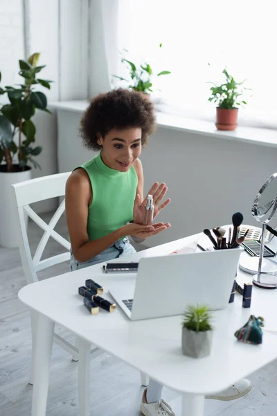 Positive african american woman holding face foundation near laptop at home — Fotografia de Stock