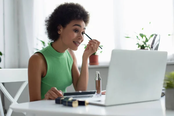 Sonriente mujer afroamericana aplicando sombra de ojos cerca de cosméticos y computadora portátil borrosa en casa - foto de stock