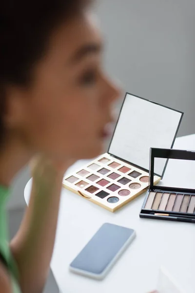Eye shadows and smartphone near blurred african american woman at home — Stock Photo