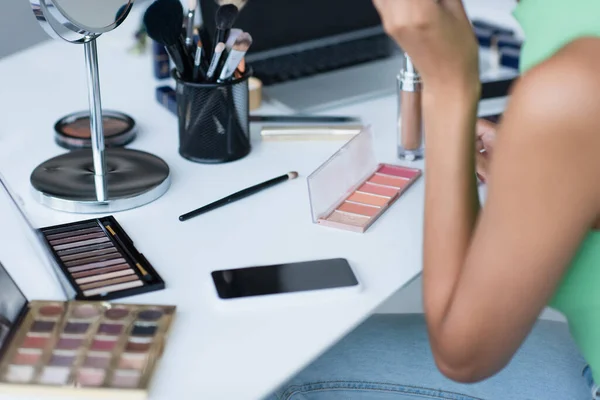 Vista recortada de espejo y cosméticos decorativos cerca del teléfono celular y la mujer afroamericana en casa - foto de stock