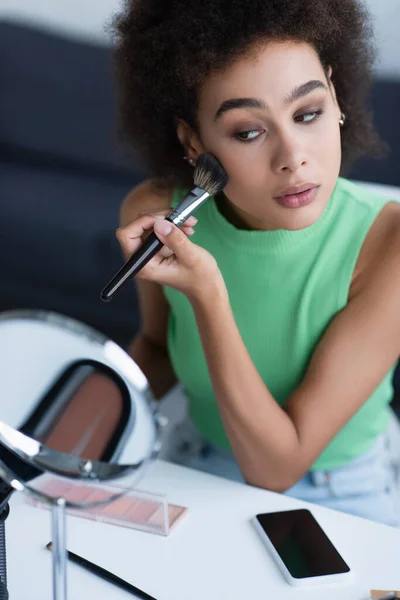 Mujer afroamericana joven aplicando rubor cerca de teléfono inteligente y espejo en casa - foto de stock