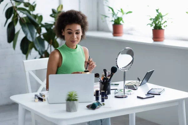 Afroamerikanerin hält Kosmetikpinsel in der Hand und schaut zu Hause auf Laptop neben Kosmetik — Stockfoto