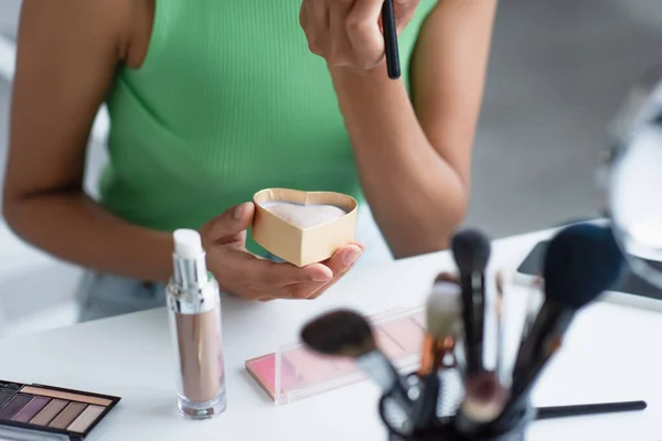 Vista recortada de mujer afroamericana sosteniendo bronceador cerca de pinceles cosméticos borrosos y teléfono inteligente - foto de stock