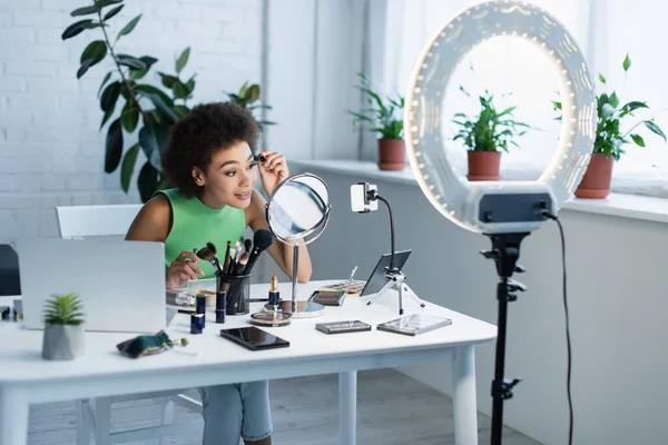 Blogueur afro-américain appliquant mascara près de gadgets et la lumière anneau à la maison — Photo de stock